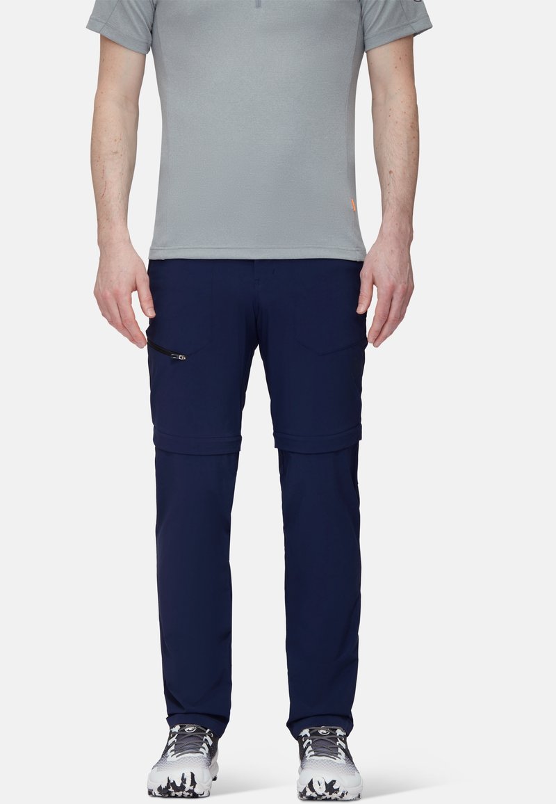 Man i grå kortärmad skjorta, marinblå byxor med avtagbara ben och vita vandringsskor, som står mot en enfärgad vit bakgrund.