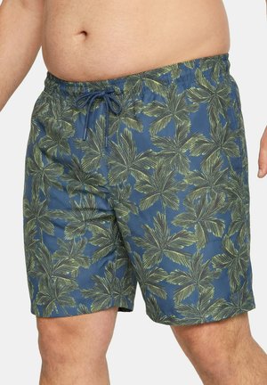 Mand iført blå badeshorts med grønne palmebladmønstre og justerbar snøre i taljen.