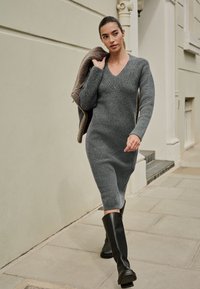 Robe en maille côtelée grise avec un col en V, des manches longues et une longueur mi-mollet, associée à des bottes noires montantes jusqu'aux genoux et portant une veste marron.