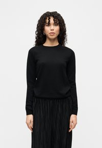 Svart strikket genser med lange ermer og rund hals, kombinert med et plissért svart skjørt. Glatt tekstur, minimalistisk design, ensfarget.