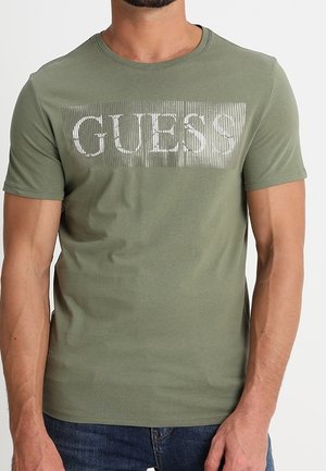 Mężczyzna ubrany w oliwkowo-zieloną koszulkę z krótkim rękawem z białym pasiastym logo „GUESS” na piersi, w parze z niebieskimi dżinsami.