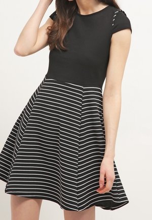 Žena v černých šatech s jednobarevným vrškem a rozšířenou sukní s bílými horizontálními pruhy, pózující s jednou rukou u hlavy.