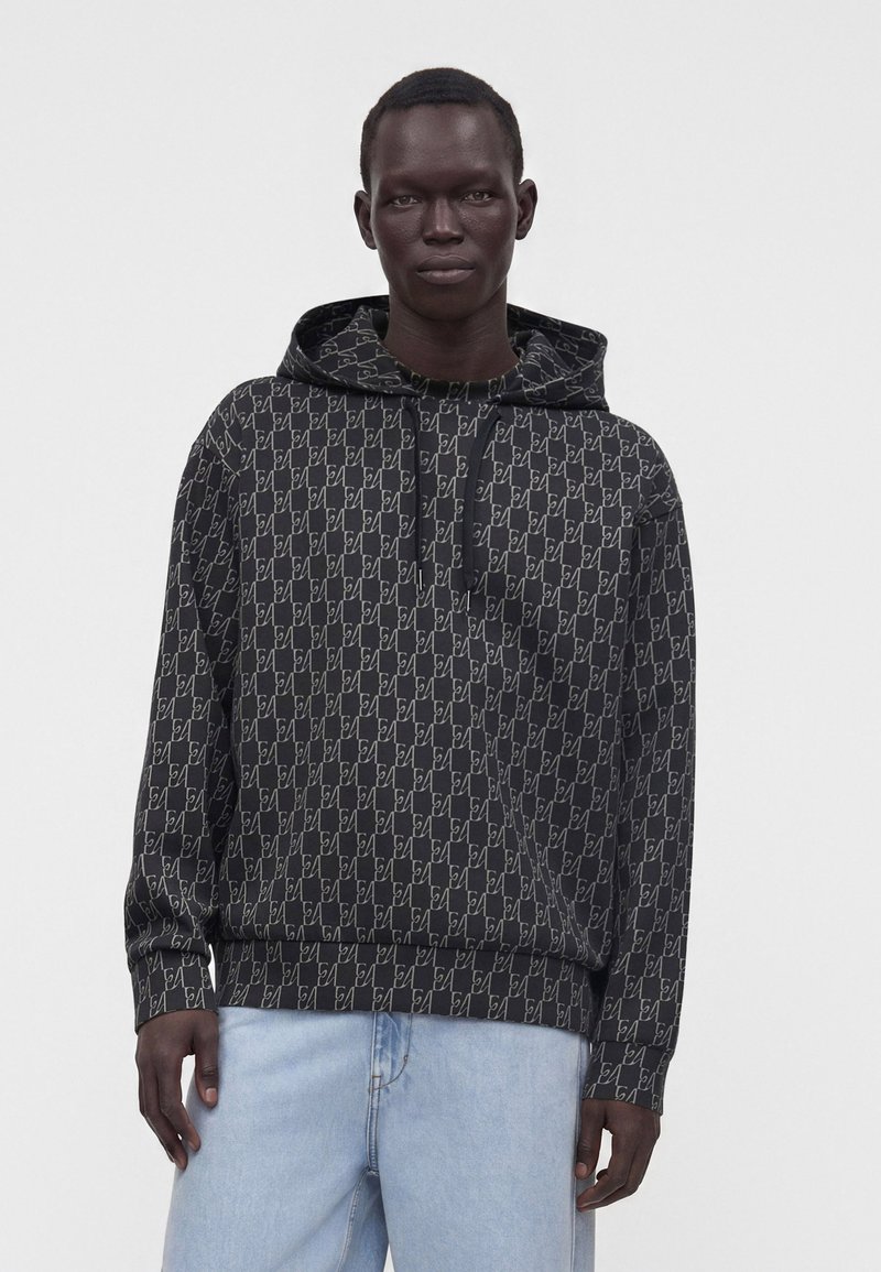 Homme portant un sweat à capuche noir avec un motif géométrique blanc répété et un jean bleu clair, debout devant un fond uni.
