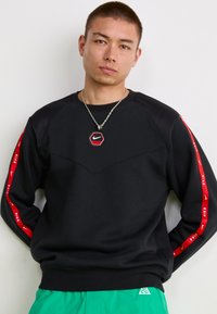 Sort sweatshirt med røde Nike-logo accenter på ærmerne, hexagonal logo vedhæng, ribbede manchetter og en tekstureret frontpanel.