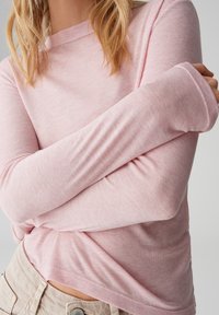 Hellrosa langärmeliges Oberteil aus weichem, strukturiertem Stoff mit Rundhalsausschnitt und verlängerten Bündchen, kombiniert mit beigen Hosen.