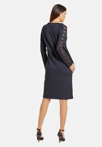 Marine jurk met lange mouwen met kantdetails op de mouwen. Strakke silhouet, knielange snit, gecombineerd met zwarte hoge hakken.