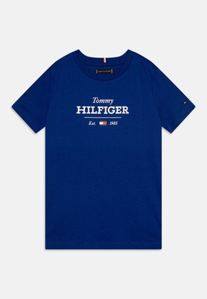 Blå T-shirt med korte ærmer og rund hals, med hvid tekst "Tommy Hilfiger Est. 1985" og lille rød, hvid og blå flaglogo på brystet og ærmet.