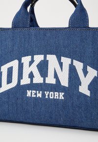 Sac fourre-tout en denim avec "DKNY NEW YORK" brodé en blanc, de forme structurée et en tissu bleu texturé. Poignées noires en haut.
