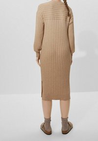 Abito maglione in beige chiaro con texture a coste, maniche lunghe, spacchi laterali e lunghezza a metà polpaccio, abbinato a scarpe marroni senza lacci e calze grigie.