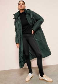 Dlhý zelený puffa kabát s kapucňou, zapínanie na zips a gombík, skombinovaný s čiernymi legínami a béžovými teniskami. Hladký materiál so šitými panelmi.