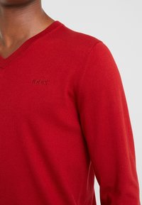 Czerwony sweter z dekoltem w serek, wykonany z miękkiego dzianiny; posiada tonowy logo na piersi oraz ściągacze przy rękawach, co gwarantuje dopasowany wygląd.