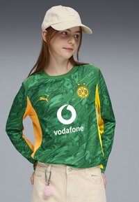 Nuori henkilö, joka pukeutuu vihreä-keltaiseen Borussia Dortmundin jalkapalloasuunsa, jossa on Vodafone-logo, beigeihin housuihin, beigeen lippalakkiin ja vaaleanpunaiseen pom-pom-avainnauhaan.