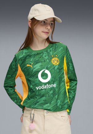 Giovane che indossa una maglietta da calcio verde e gialla del Borussia Dortmund con il logo Vodafone, pantaloni beige, cappellino beige e portachiavi con pompon rosa.