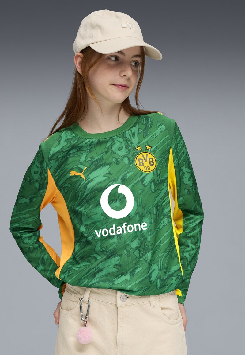 Nuori henkilö, joka pukeutuu vihreä-keltaiseen Borussia Dortmundin jalkapalloasuunsa, jossa on Vodafone-logo, beigeihin housuihin, beigeen lippalakkiin ja vaaleanpunaiseen pom-pom-avainnauhaan.