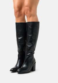 Bottes en cuir noir montant jusqu'aux genoux, avec une texture lisse, des bouts carrés et des talons épais. Elles comportent une fermeture éclair sur le côté intérieur.