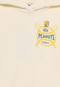 Ein weißer Hoodie mit einem grünen und gelben Grafikdesign eines Baseballs, auf dem der Text "PEANUTS" und "1950" steht, gekrönt von einem Comic-Vogel, der einen Hut trägt.