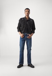 Jachetă din denim neagră cu accente uzate, tricou negru și blugi drepți albaștri. Adidași negri completează ținuta. Fundal alb.