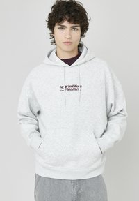 Sweat à capuche gris avec une poche avant, un cordon de serrage et un texte brodé en bourgogne. Tissu doux, coupe ample et poignets côtelés aux manches.