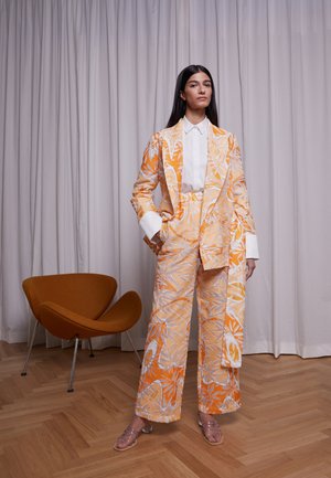 Fato com padrão floral em laranja e creme, com calças de perna larga, combinado com uma camisa branca e sandálias transparentes de tiras. Tecido texturizado.
