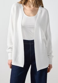 Vit kofta med öppen front, ribbad textur och långa ärmar, tillsammans med en åtsittande vit topp och mörkblå jeans.