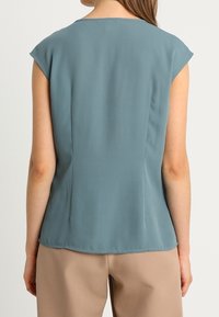 Blouse bleu clair à manches courtes avec une texture lisse et fluide ; coupe ajustée avec des coutures au dos et un col arrondi.