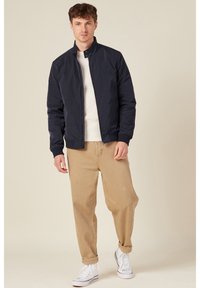 Marine-blau Bomberjacke aus glattem Stoff, mit Stehkragen und Reißverschluss, kombiniert mit beigen Hosen und weißen Sneakers.