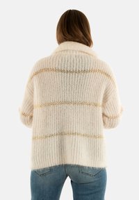 Frau, die sich abwendung zeigt, trägt einen lockeren, cremefarbenen Strickpullover mit goldenen horizontalen Streifen und blaue Jeans vor einem einfarbigen Hintergrund.