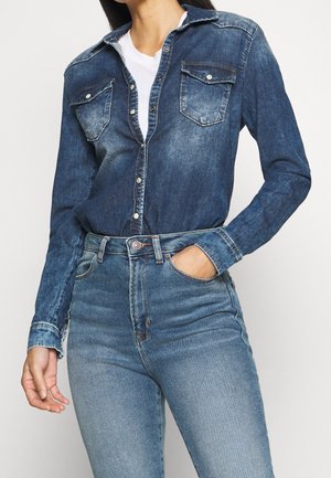 Kobieta w ciemnym dżinsowym koszuli na guziki z kieszeniami z klapami z przodu, nosząca białą koszulkę pod spodem, w połączeniu z jasnoniebieskimi dżinsami z wysokim stanem.