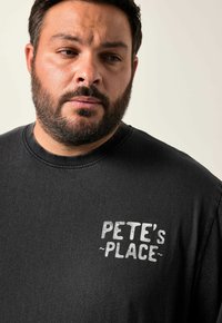 Svart bomull t-shirt med rund halsringning, texturerad tyg och en vit tryckt logotyp "PETE'S PLACE" på bröstet.