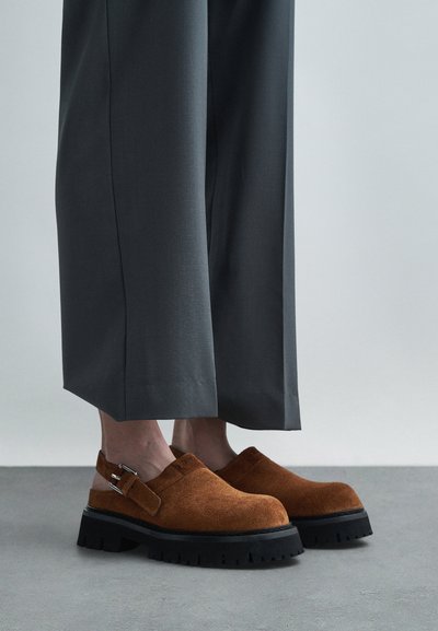 Sabots en daim marron avec une semelle noire épaisse, dotés d'une boucle sur le côté, assortis à un pantalon large gris. Dessus plat et texturé.