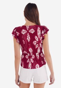 Bordeauxfarbenes Oberteil mit weißen Blumenmustern, kurzen Rüschensäumen, geraffter Taille und einem strukturierten Stoff. Kombiniert mit weißen Shorts.