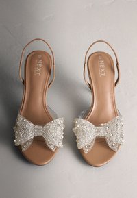 Sandales ouvertes couleur beige avec brides à la cheville et grands nœuds blancs perlés sur le devant, taille 6½/40, marque "NEXT" visible à l'intérieur.