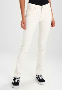 Jeans blancs slim en denim, avec fermeture à bouton et zip, cinq poches et une texture lisse. Portés avec des baskets noires.