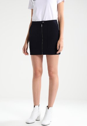 Femme portant une mini-jupe noire avec une fermeture éclair à l'avant et des poches, un t-shirt blanc et des bottines blanches sur un fond uni.