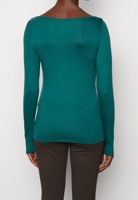 Långärmad topp i teal, tillverkad av mjukt, elastiskt tyg. Har en vid halsringning och en figurnära siluett. Bärs med mörkbruna byxor.