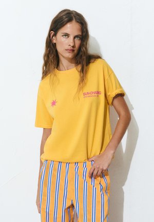 Femeie cu păr lung și șaten, purtând un tricou galben cu text roz și pantaloni portocalii cu dungi, stând lângă un perete alb.