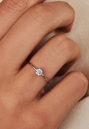 Silberner Ring mit einem runden, klaren Zirkonia-Hauptstein auf einem schlanken Band, das an einer Hand mit strukturiertem Haut getragen wird.