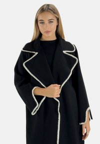 Black coat with a shawl collar and white trim, featuring a textured fabric and open front design. Long sleeves with decorative accents.