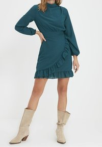 Teealfarbenes Kleid mit strukturierten Punkten, langen Ärmeln, schwingendem Saum und tailliertem Schnitt. Kombiniert mit beigen, lässigen Stiefeln mit Absatz.