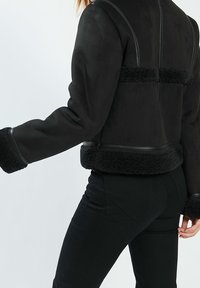 Chaqueta negra con una textura suave y peluda, que presenta acentos de cuero. El diseño incluye un corte corto y puños anchos y esponjosos.