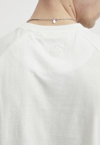 Hvit bomull T-skjorte med rund hals og raglanermer. Har et subtilt preget design på baksiden og en teksturert overflate.
