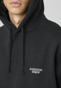 Hoodie noir avec une coupe décontractée, dotée d'une poche kangourou à l'avant, de cordons de serrage et du texte "POTENTIAL INSIDE" en broderie blanche.