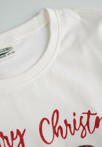 Biała bawełniana koszulka z okrągłym dekoltem, krótkimi rękawami i czerwonym brokatowym napisem "Wesołych Świąt" z przodu.