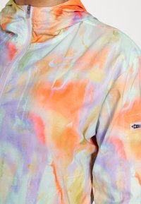 Nike větrovka s lehkým, prodyšným materiálem. Obsahuje vícebarevný abstraktní vzor v odstínech oranžové, fialové a zelené.