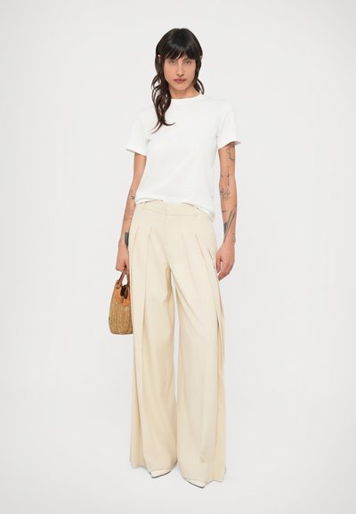 Biele tričko spojené s béžovými nohavicami s vysokým pásom a širokými nohavicami s záhybmi. Drží pletenú kabelku s hnedým akcentom.