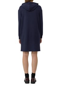 Frau in einem navyblauen, kapuzenförmigen, langärmeligen Kleid, schwarzen Socken und dunkelroten Plateauschuhen, die mit dem Rücken zur Kamera auf einem weißen Hintergrund steht.