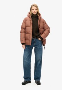 Veste matelassée marron avec capuche, présentant un design matelassé, portée sur un pull en maille sombre et un jean large bleu, associée à des chaussures noires.