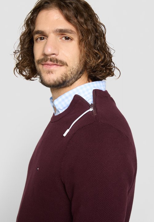 Bordo pulover s teksturiranim pletenjem, s ovratnikom na patentni zatvarač i bijelim akcentima, složen preko svijetloplavog košulje s kariranim uzorkom.