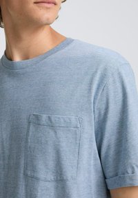 Tricou din bumbac albastru, cu mâneci scurte, de tip guler rotund și un buzunar cu petic pe partea stângă a pieptului, având o textură subtilă în dungi.
