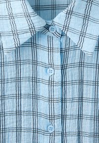 Chemise bleu clair boutonnée avec motif à carreaux noir et blanc et col pointu, vue en gros plan sur les boutons avant et la texture du tissu.